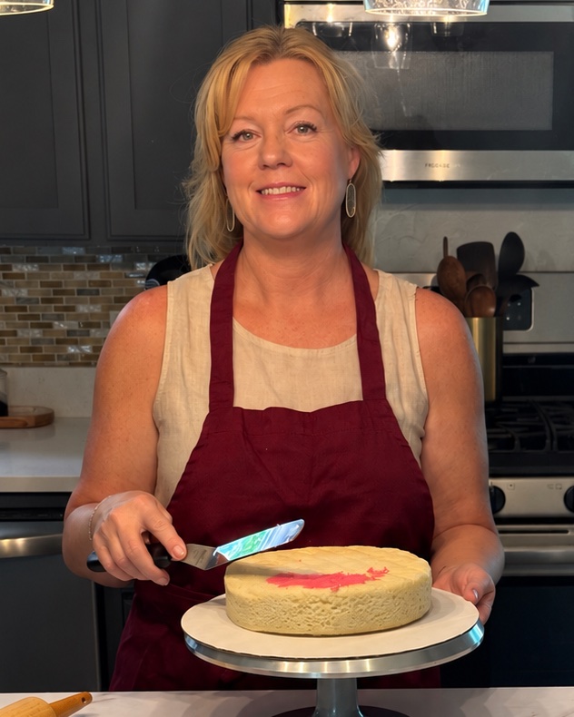 Lisa smiling in her kitchen in a burgundy apron, frosting a cake on a cake stand
